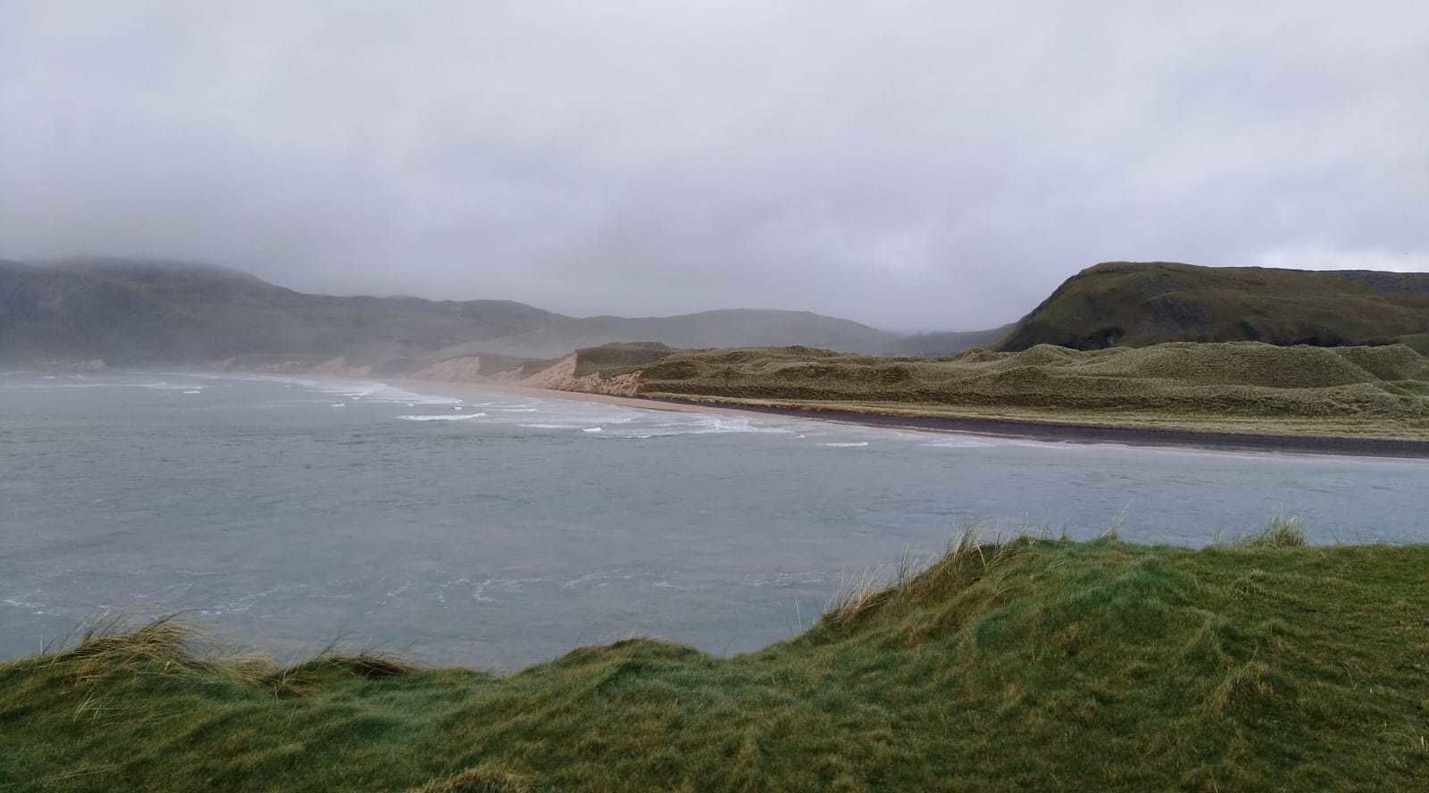 Paisagem costeira com praia, dunas gramadas e céu nublado.