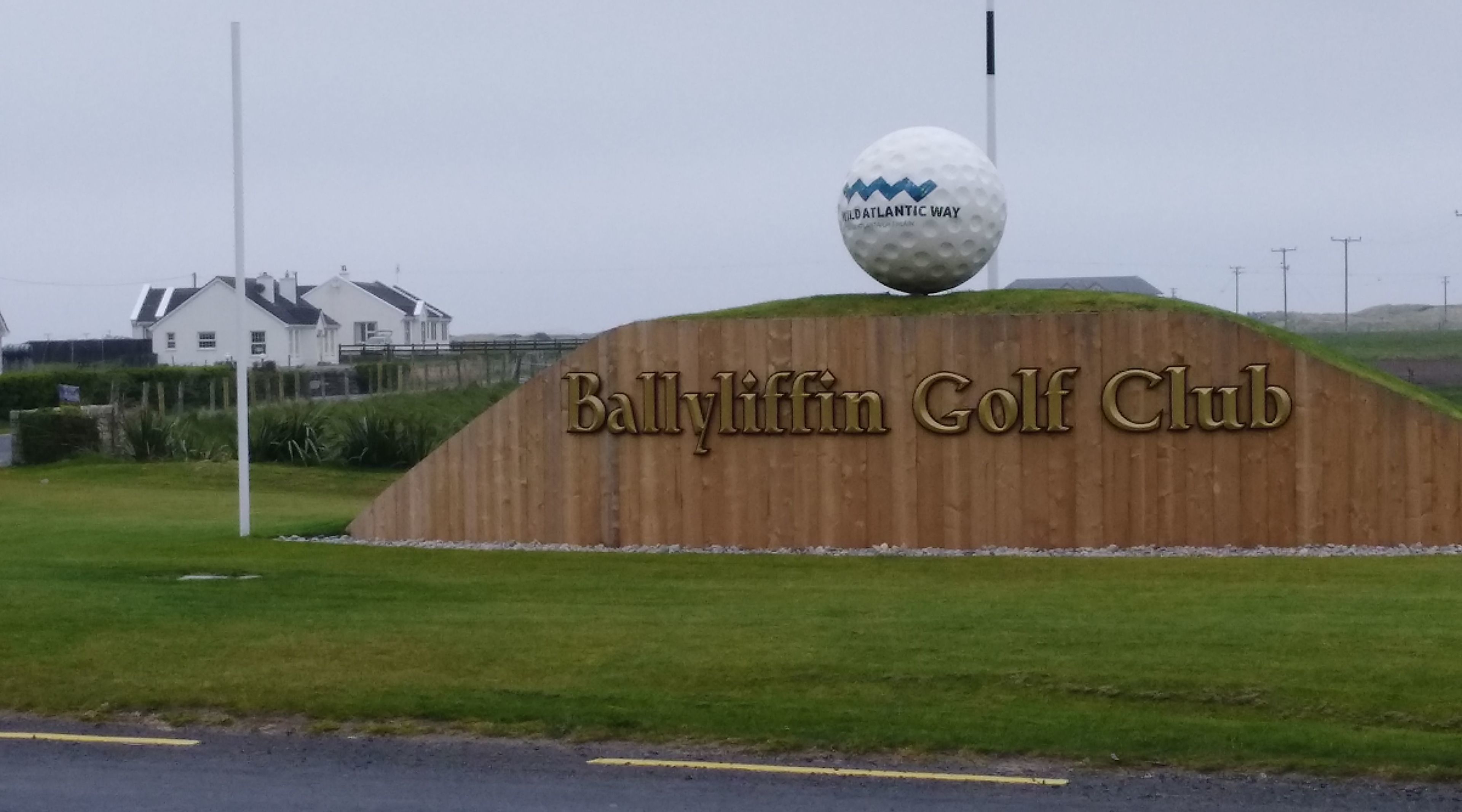 A golf club sign with a golf ball on top, surrounded by a grassy area and a wooden structure.