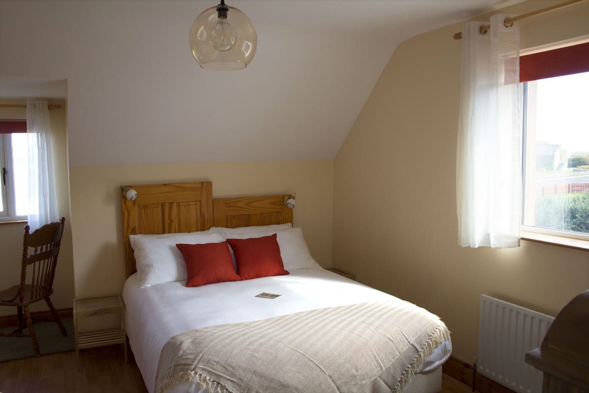 A bedroom with a wooden headboard, a bed with white and red bedding, a bedside table, and a window with curtains.