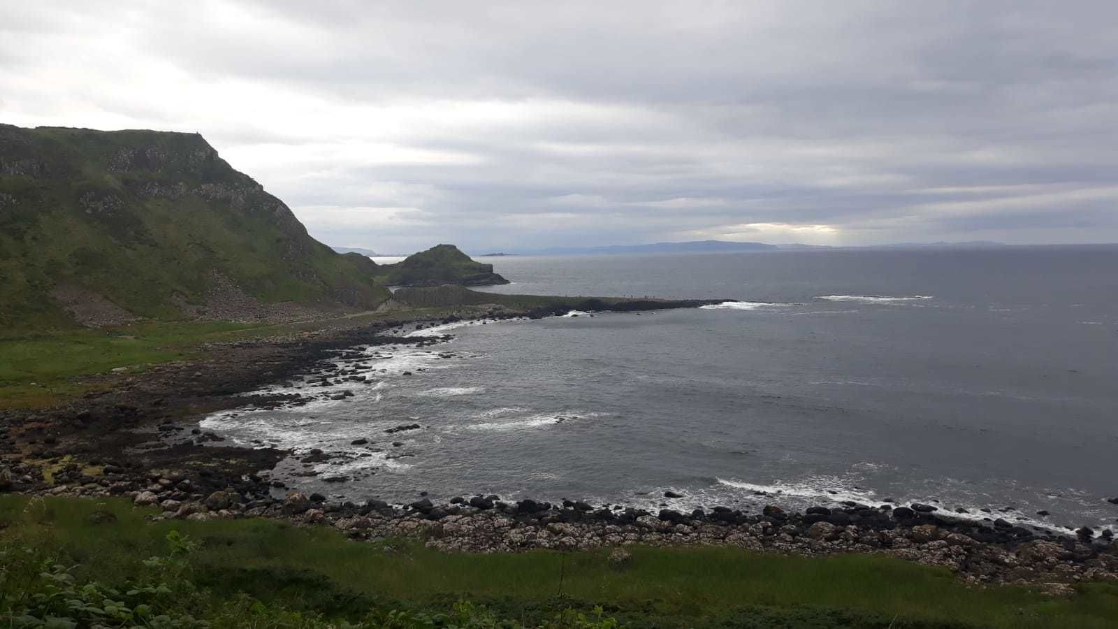 A coastal landscape with rocky shores, grassy areas, and a distant view of the sea under a cloudy sky.