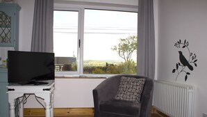 Una sala de estar con un televisor sobre una mesa blanca, un sofá gris y vistas al campo a través de una ventana.