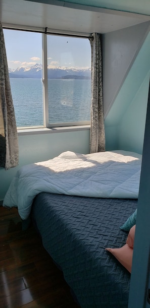 A room with a bed, a window with curtains, and a view of mountains and water.