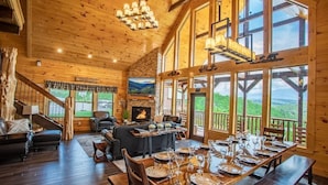 Ein geräumiges Holzhaus mit Essbereich, gedecktem Tisch, Kamin und großem Fenster mit Blick auf eine Berglandschaft.