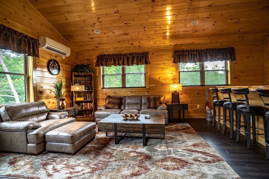 A cozy cabin interior with a wooden ceiling, leather furniture, a coffee table, and a bar area with stools.