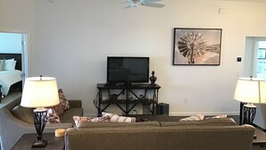 Ein Wohnzimmer mit Deckenventilator, Fernseher auf einem Schrank, zwei Lampen, einem gerahmten Bild und einem Sofa.