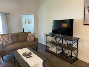 Un salon comprenant un canapé, une table basse, un téléviseur posé sur un meuble et une petite étagère avec quelques objets.
