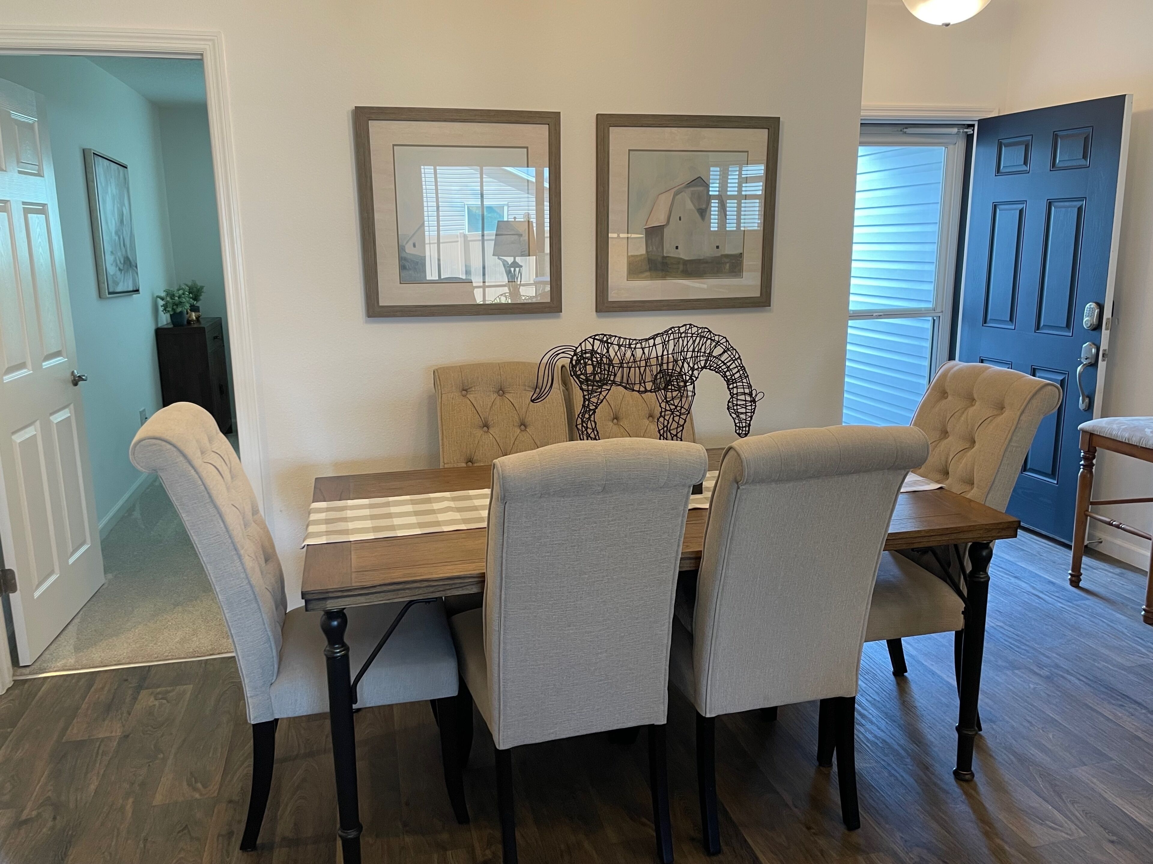A dining area with a wooden table and four upholstered chairs, a framed picture on the wall, and a blue door in the background.