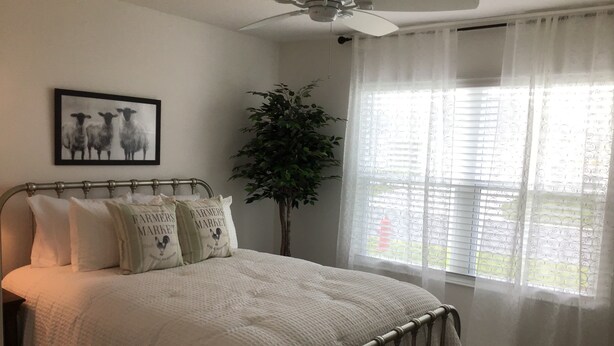 Ein Schlafzimmer mit einem Bett, einem Deckenventilator, einem Fenster mit Vorhängen, einer Pflanze und einem gerahmten Bild an der Wand.