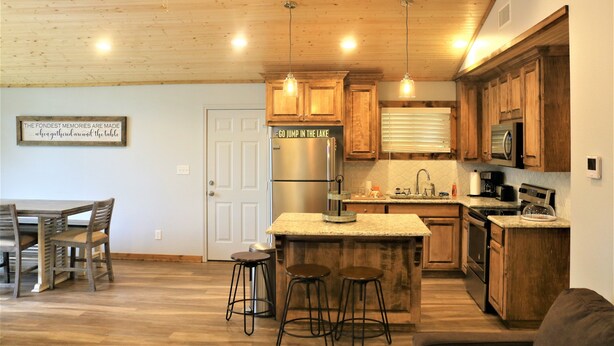 Una cocina moderna con armarios de madera, un refrigerador de acero inoxidable y una encimera de granito. Hay dos taburetes de bar y una mesa de comedor con sillas.