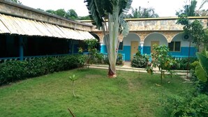 A courtyard with a building featuring a stone facade and blue accents, surrounded by greenery and a palm tree. - Orchidée Hôtel (Kpalimé)