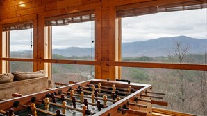 A room with a foosball table and a sofa, large windows with a mountain view.