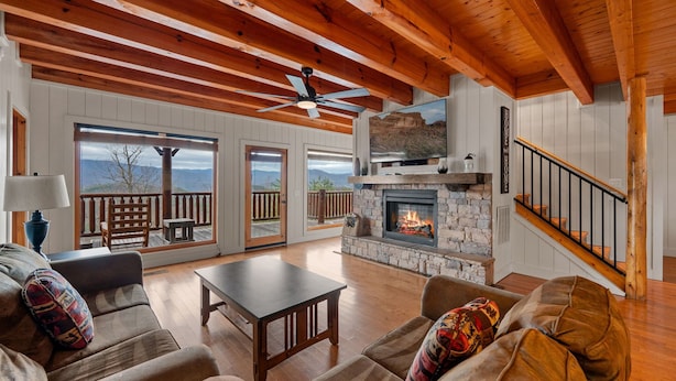 A spacious living room with a stone fireplace, wooden ceiling beams, and large windows offering a view of the outdoors.