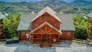 A log cabin with a front porch, surrounded by a forested mountain backdrop.
