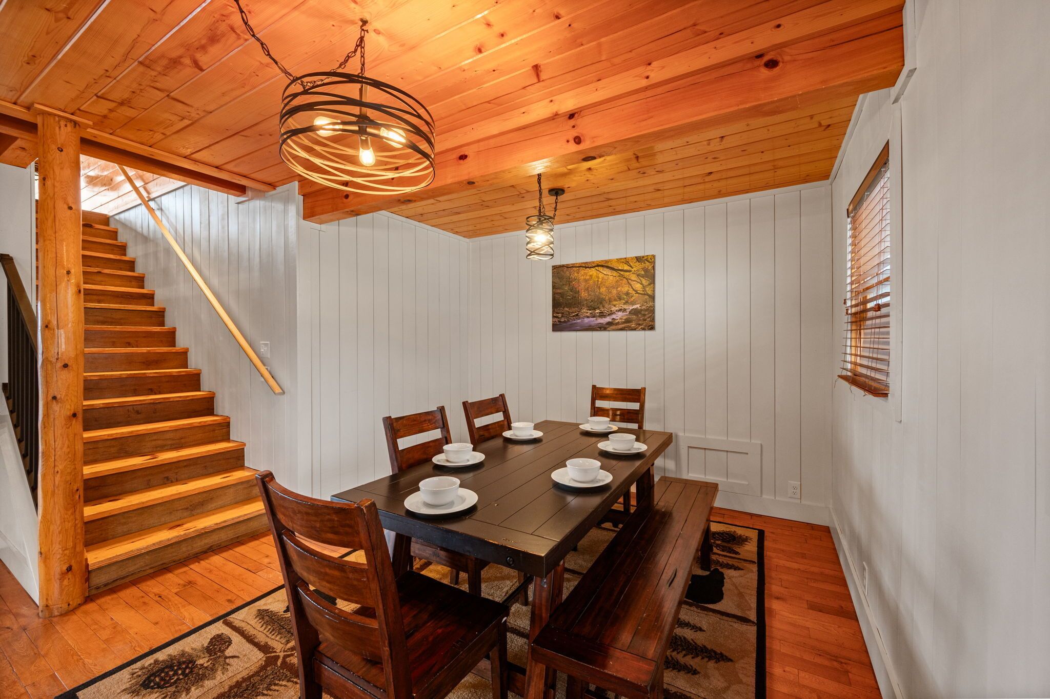 A dining area with a wooden table set for six, a staircase, and a painting on the wall.