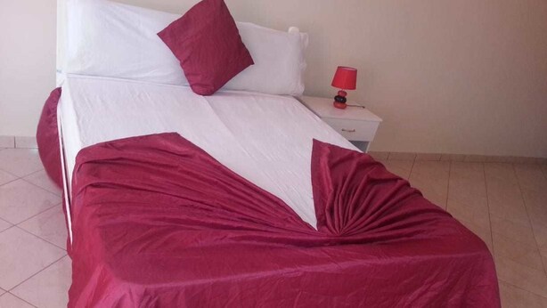 A bed with a white and red bedspread, a red pillow, and a small bedside table with a red lamp. - Casa anilda & albino (São Filipe)
