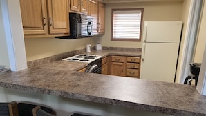 Una cocina con encimeras de granito, armarios de madera, un microondas negro, un refrigerador blanco y una ventana con persianas.