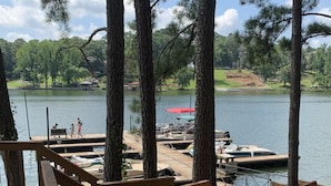 Un muelle de madera con barandilla, con vistas a un lago tranquilo con barcos y un área de césped al fondo.