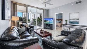 Een moderne woonkamer met een leren bank, een salontafel en een televisie op een standaard. Er is een balkon met uitzicht op de stad en een plafondventilator.