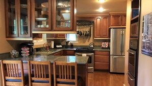 A kitchen with wooden cabinets, a stainless steel refrigerator, and a central island with chairs. - Spacious Talkeetna home on Ruth Glacier Avenue (Talkeetna)