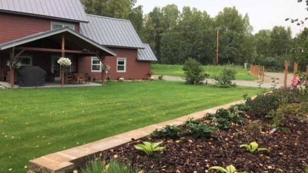 A red building with a covered porch, a lawn, and a garden area with mulch and plants. - Spacious Talkeetna home on Ruth Glacier Avenue (Talkeetna)