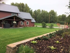 A red building with a covered porch, a lawn, and a garden area with mulch and plants. - Spacious Talkeetna home on Ruth Glacier Avenue (Talkeetna)