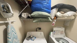 Un área de lavandería con lavadora, secadora y un estante con almohadas y una botella de detergente.