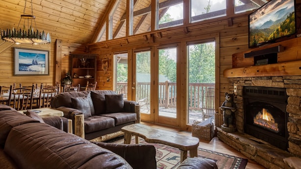 A cozy cabin interior with a stone fireplace, wooden ceiling, and large windows. - Appalachian Adventure - 4 Bedroom sleeps 14! (Gatlinburg)