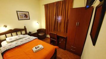 Quarto de hotel com cama, secretária, cadeira, guarda-roupa e televisão.