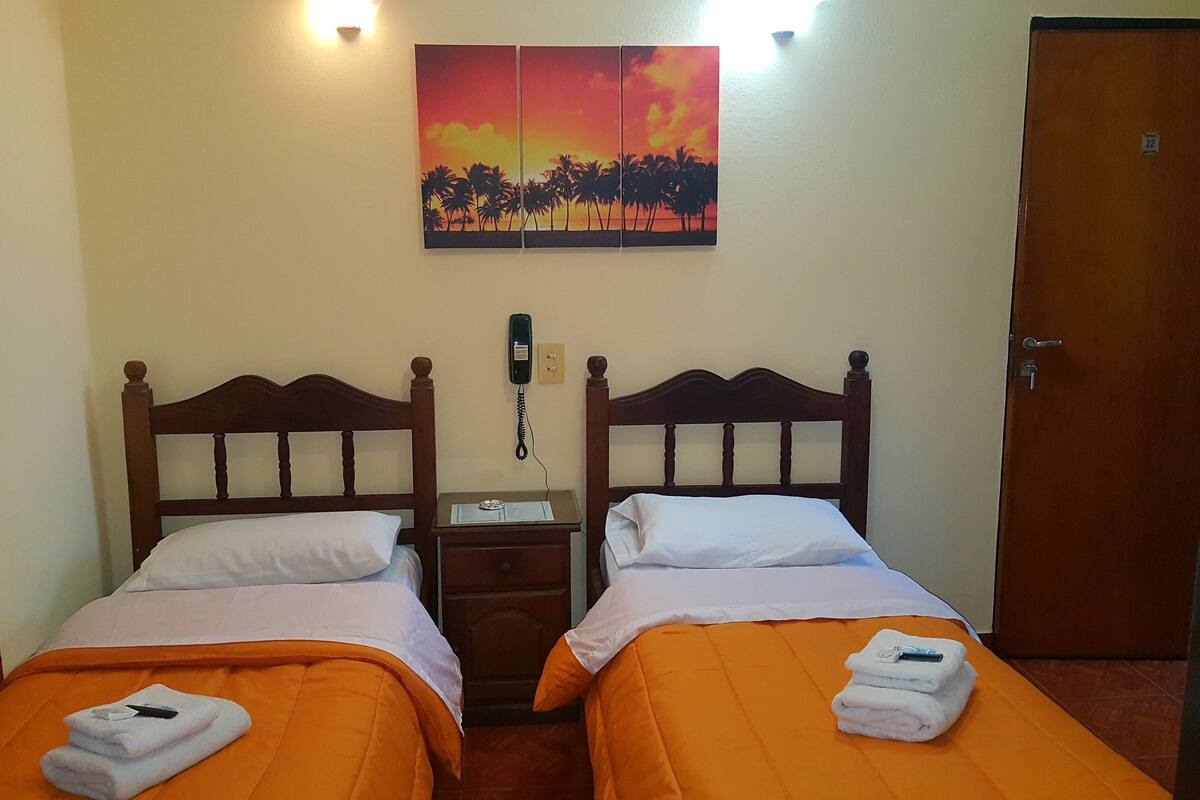 Une chambre d’hôtel avec deux lits, une couvre-lit orange, un tableau représentant un coucher de soleil, un téléphone sur la table de chevet et une porte avec un numéro.