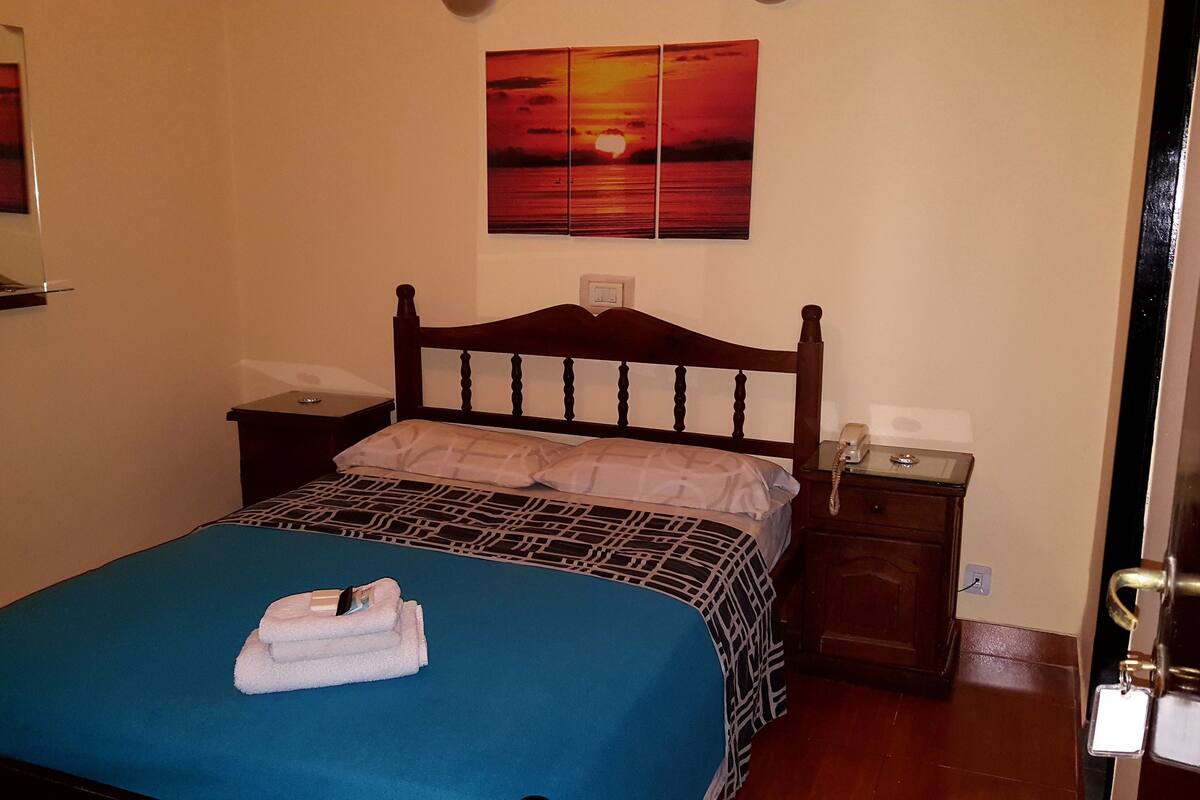 Un lit bien fait, avec une couvre-lit bleue et des oreillers blancs, une tête de lit en bois, une table de chevet avec une lampe et un tableau représentant un coucher de soleil accroché au mur.
