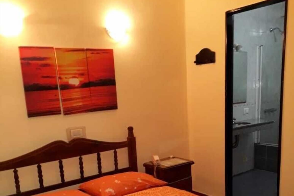 A hotel room with a bed, a nightstand, a painting, and a bathroom visible through a glass door.