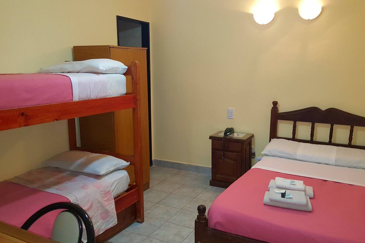 Une chambre avec des lits superposés, un lit simple, une petite commode en bois et une table de chevet avec une lampe.