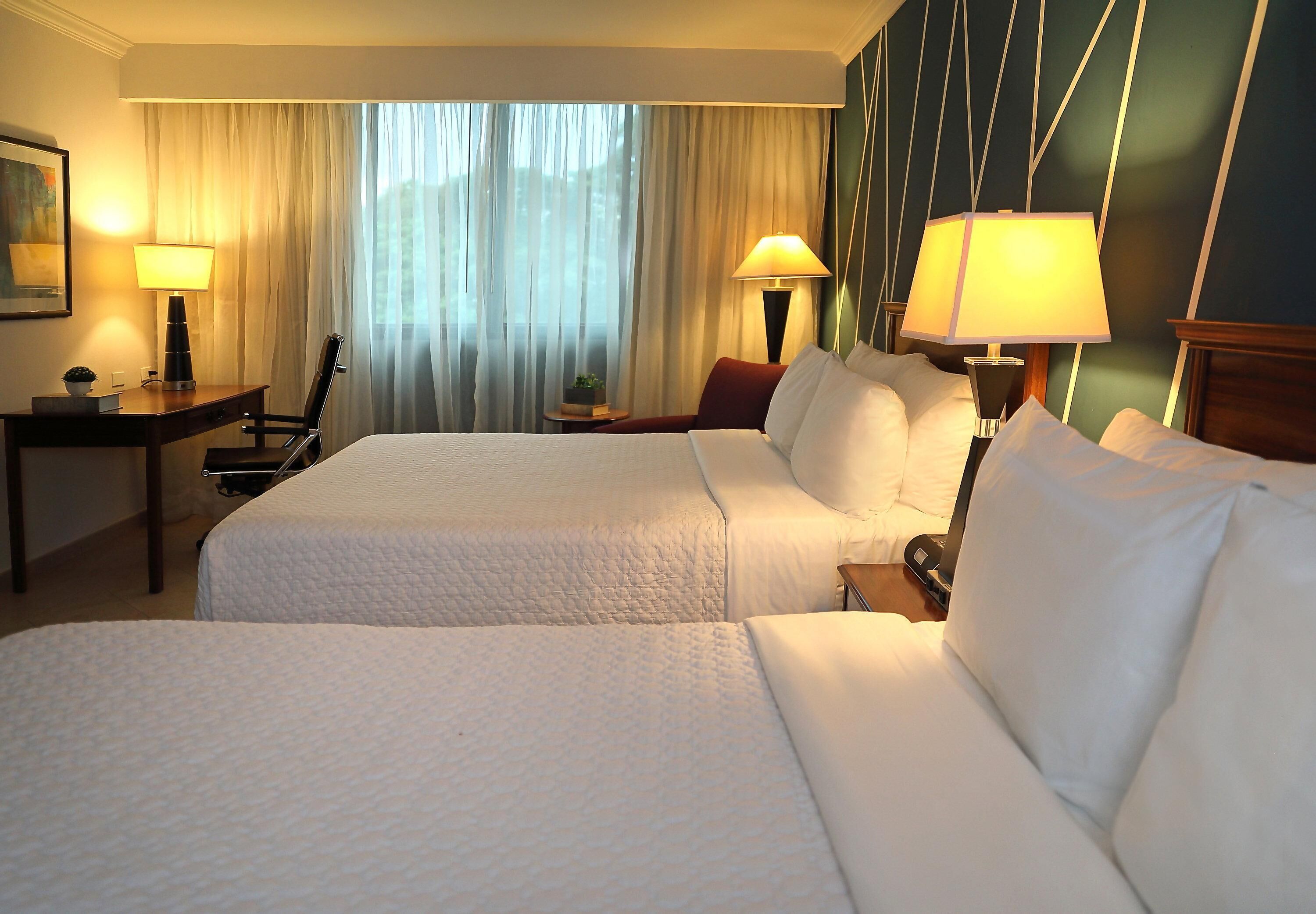 A hotel room with two beds, a desk, lamps, and a view of the outdoors through sheer curtains.