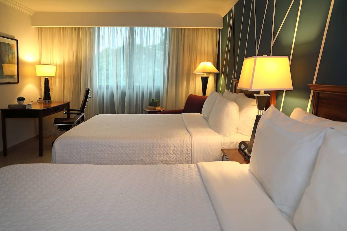 A hotel room with two beds, a desk, lamps, and a view of the outdoors through sheer curtains.