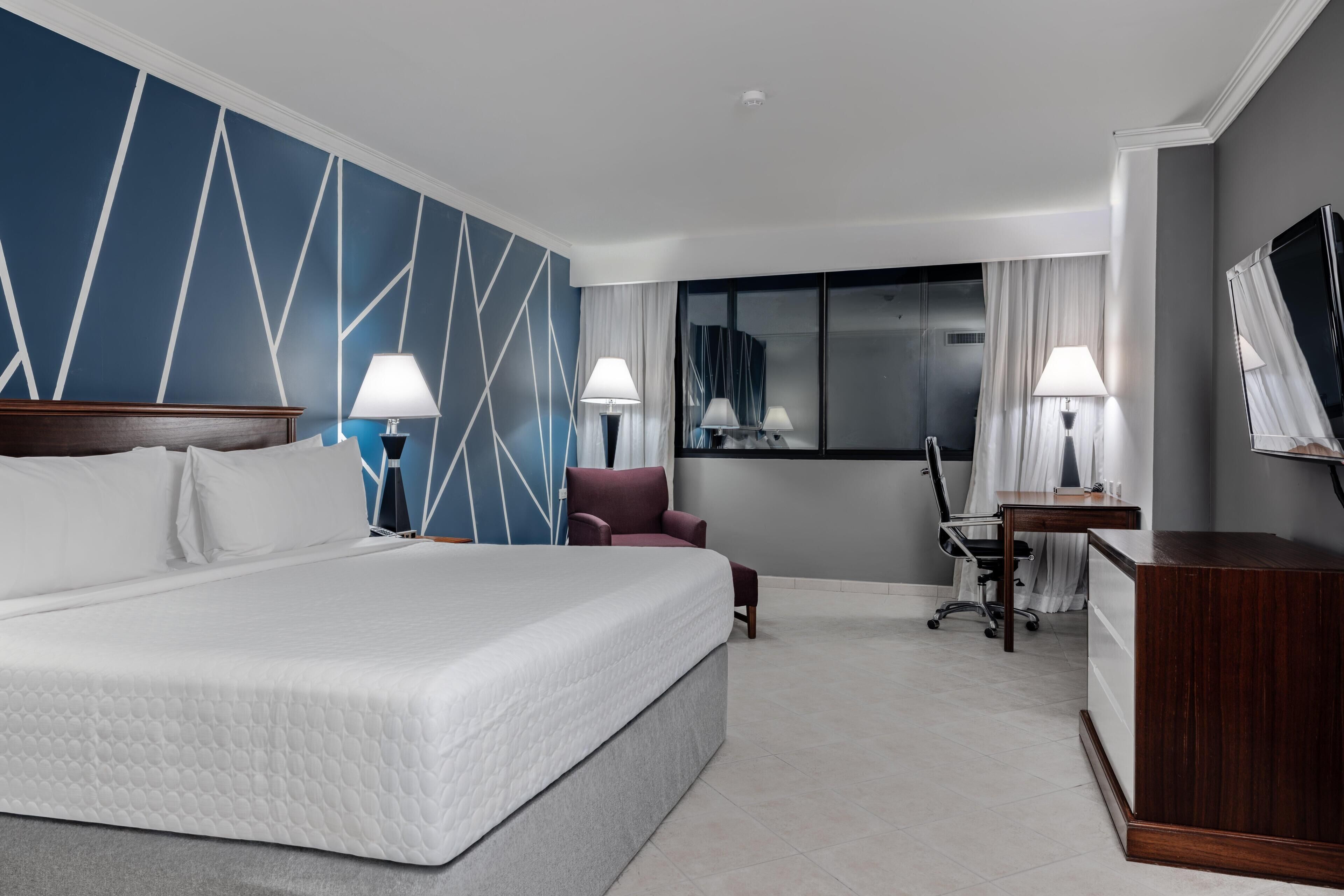 A hotel room with a large bed, two bedside tables with lamps, a desk, a chair, and a TV.