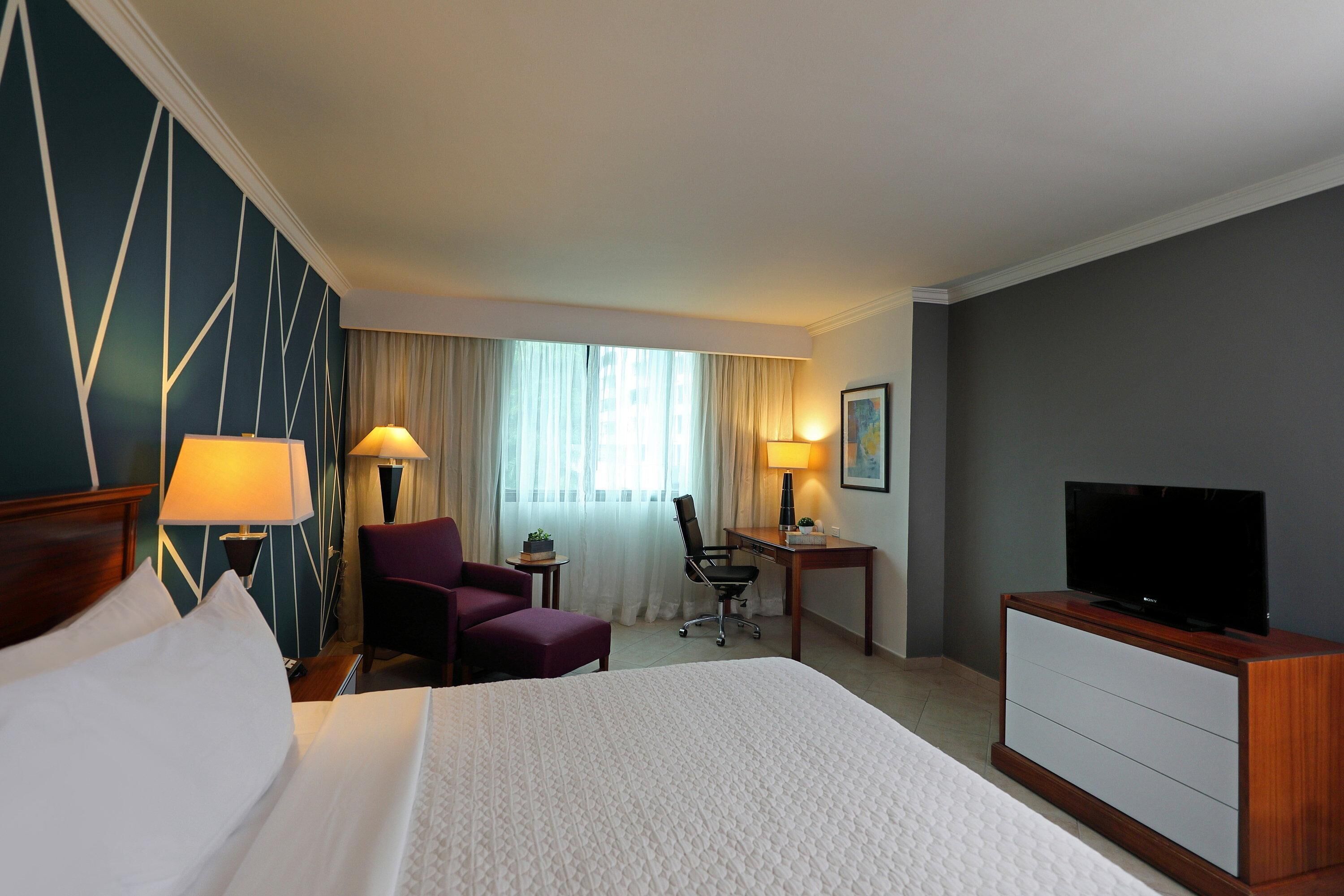 A hotel room with a large bed, a desk with a chair, a television, and a small table with a lamp.