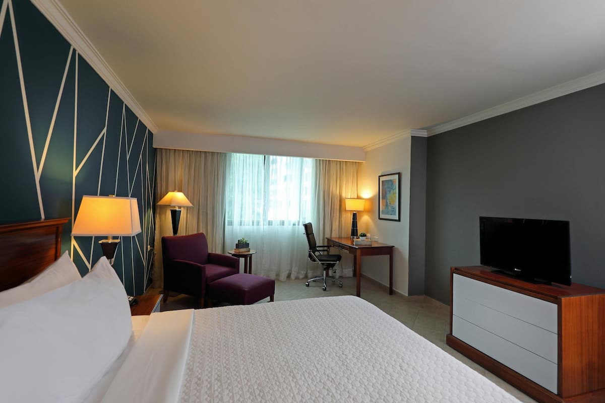 A hotel room with a large bed, a desk with a chair, a television, and a small table with a lamp.
