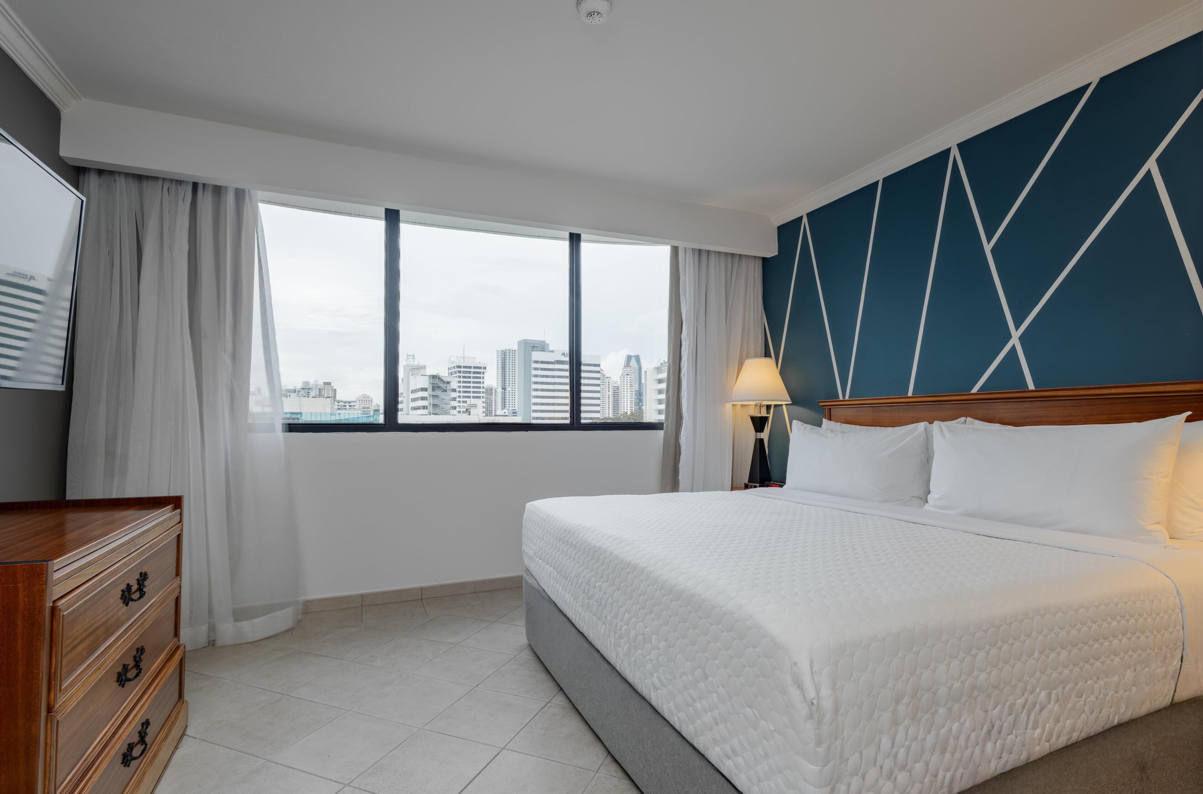 A hotel room with a large bed, a dresser, a nightstand, a lamp, and a view of the cityscape through the window.
