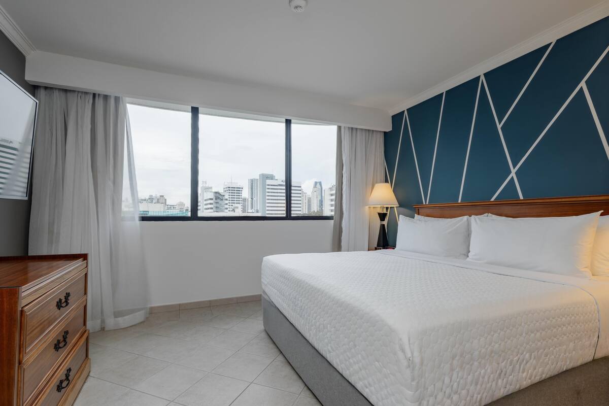 Ein Hotelzimmer mit einem großen Bett, einem Schminktisch, einem Nachttisch, einer Lampe und Blick auf die Stadtlandschaft durch das Fenster.