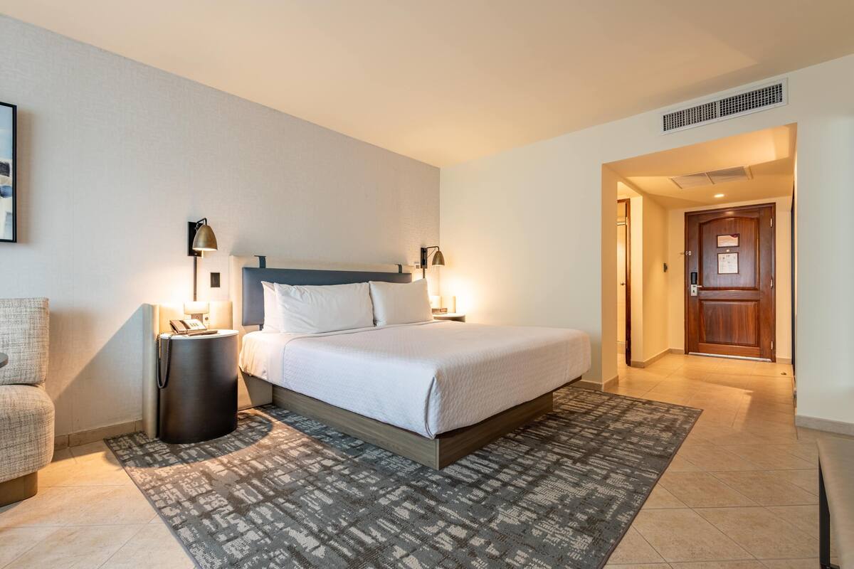 Ein Hotelzimmer mit Bett, Nachttischlampen, einem Nachttisch, einem gemusterten Teppich und einer Tür zu einem weiteren Raum.