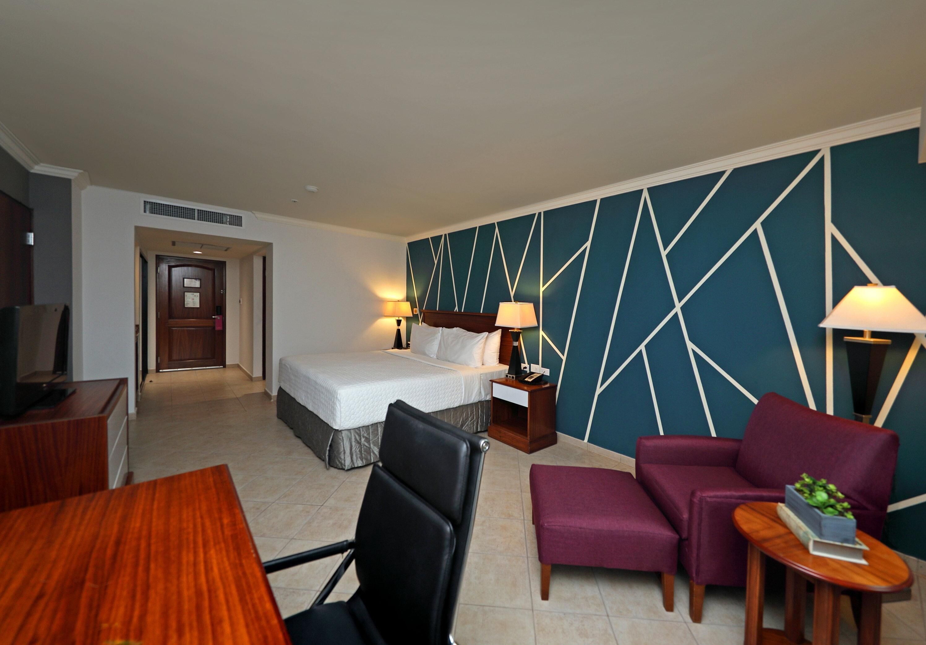 A hotel room with a bed, two bedside tables with lamps, a desk with a chair, a sofa, and a small table with a plant.