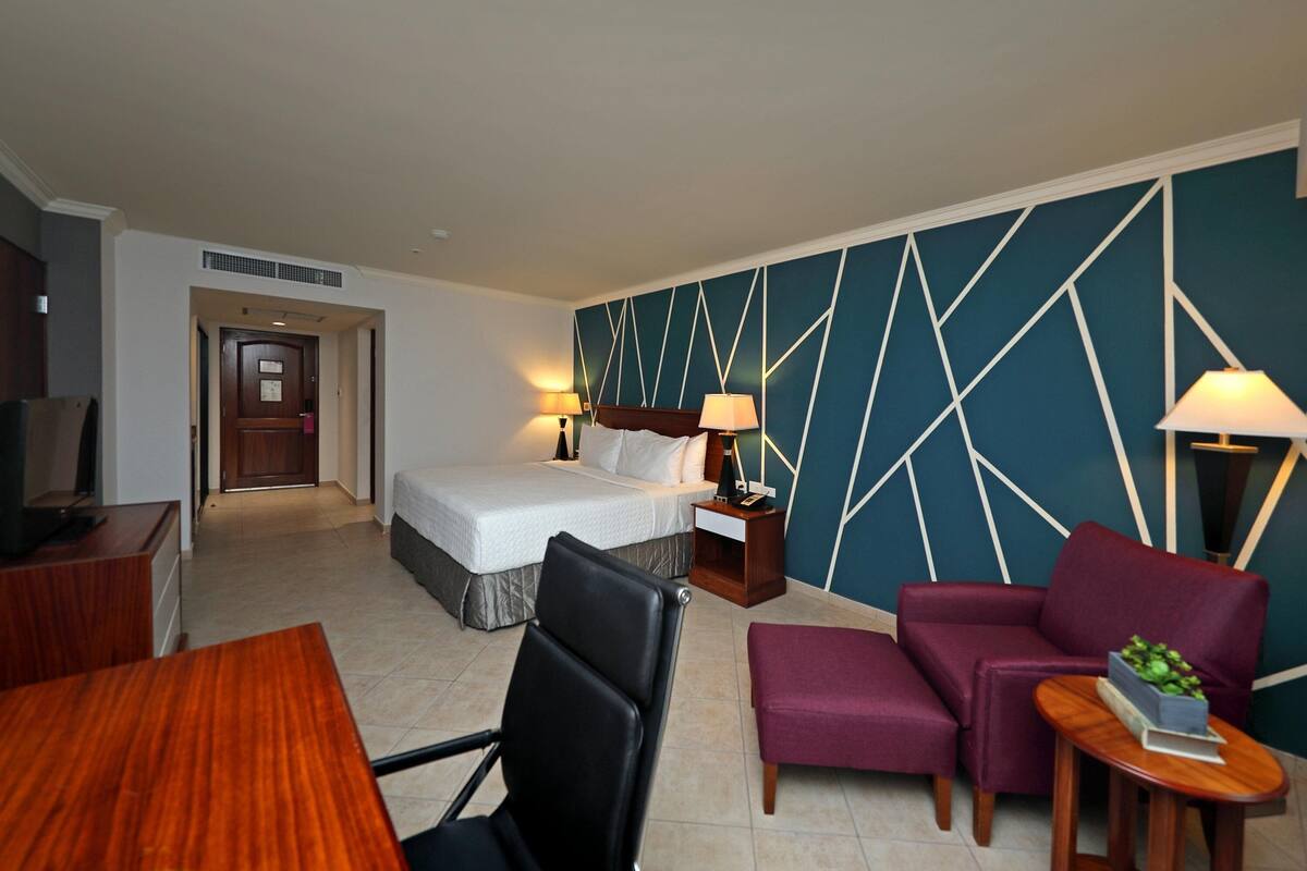 Ein Hotelzimmer mit einem Bett, zwei Nachttischen mit Lampen, einem Schreibtisch mit Stuhl, einem Sofa und einem kleinen Tisch mit Pflanze.