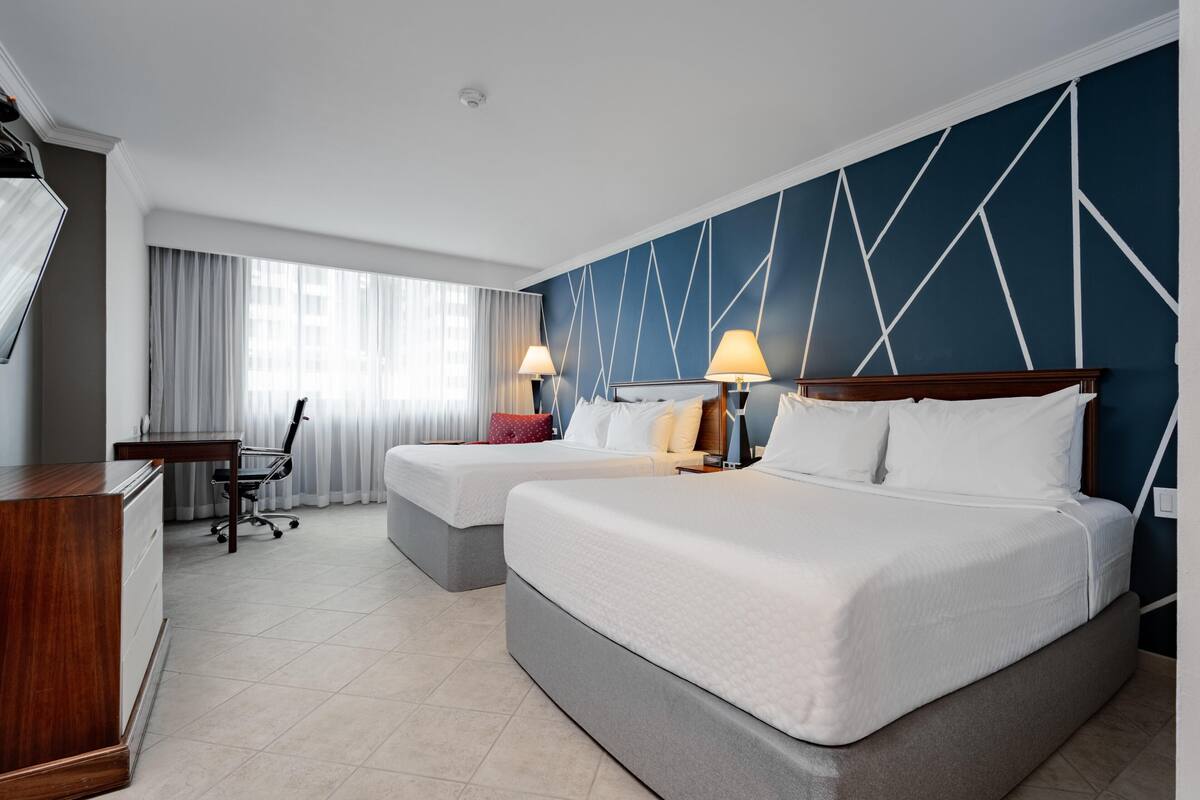 Ein Hotelzimmer mit zwei Betten, einem Schreibtisch, einem Stuhl und einem großen Fenster mit Vorhängen.