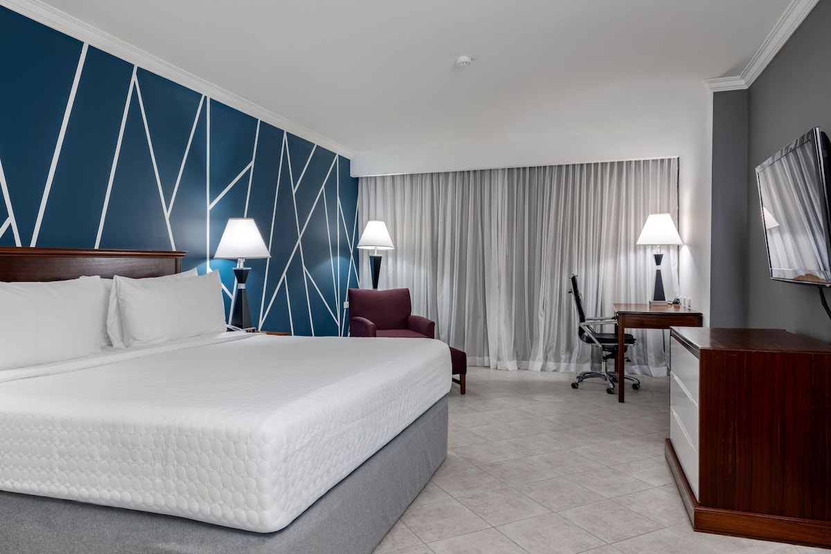 Ein Hotelzimmer mit einem großen Bett, zwei Nachttischlampen, einem Schreibtisch mit Stuhl, einem Fernseher und einem Fenster mit Vorhängen.