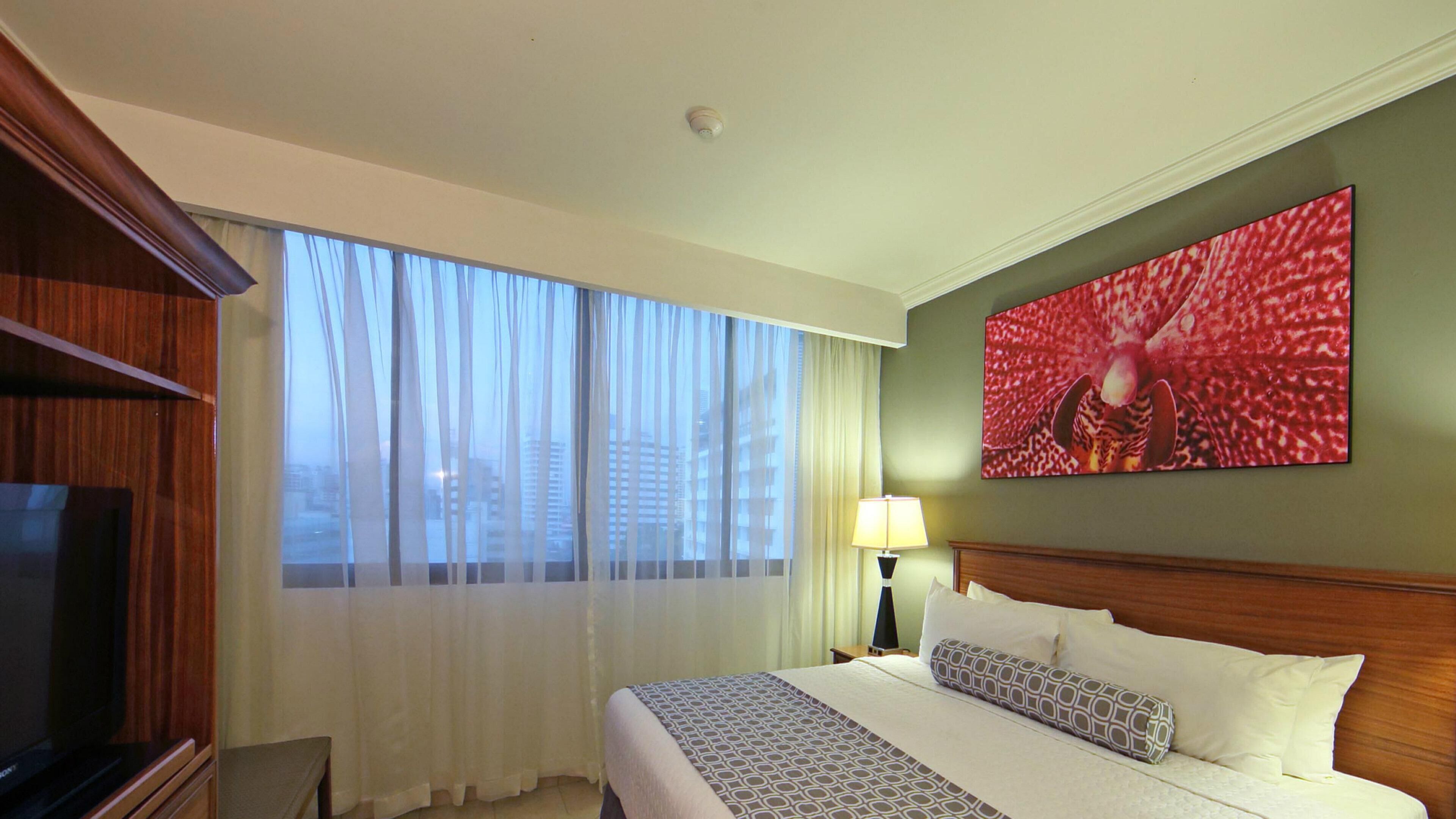 A hotel room with a large bed, a television, a painting, and a view of buildings through sheer curtains.