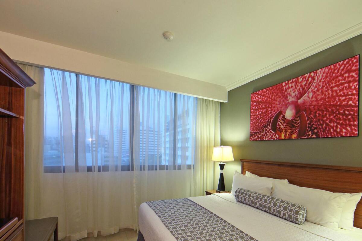 Ein Hotelzimmer mit einem großen Bett, einem Fernseher, einem Gemälde und Blick auf Gebäude durch transparente Vorhänge.
