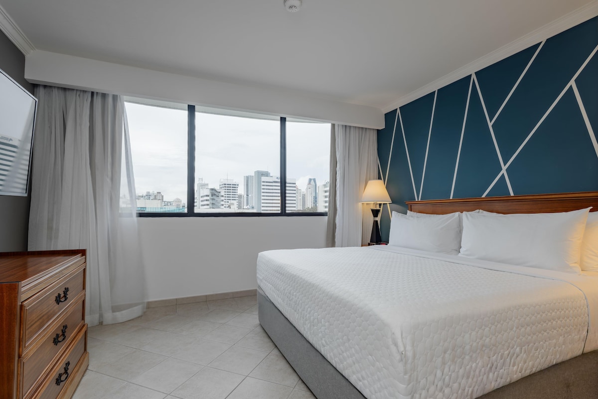 Ein Hotelzimmer mit einem großen Bett, einem Schminktisch, einem Nachttisch, einer Lampe und Blick auf die Stadtlandschaft durch das Fenster.