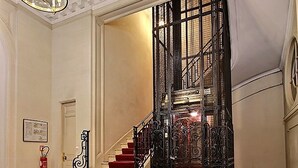 Un magnifique escalier, avec des marches recouvertes de moquette rouge et une rampe en métal noir, menant à une pièce dotée d’une grande fenêtre et d’un lustre.