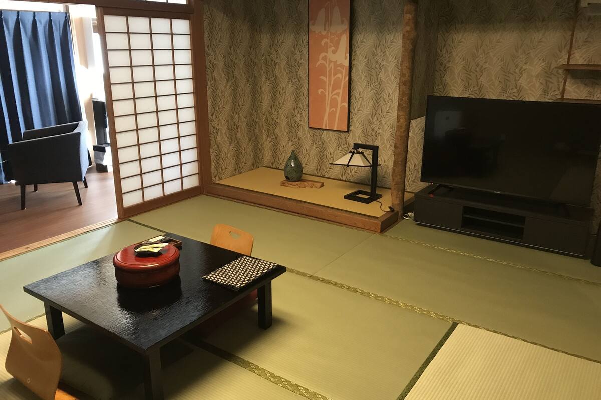 Ett traditionellt japanskt rum med tatamimattor på golvet, ett lågt bord med en röd behållare, en tv och en dekorativ väggdekoration.