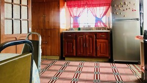 A kitchen with wooden cabinets, a refrigerator, a window with a red curtain, and a tiled floor.